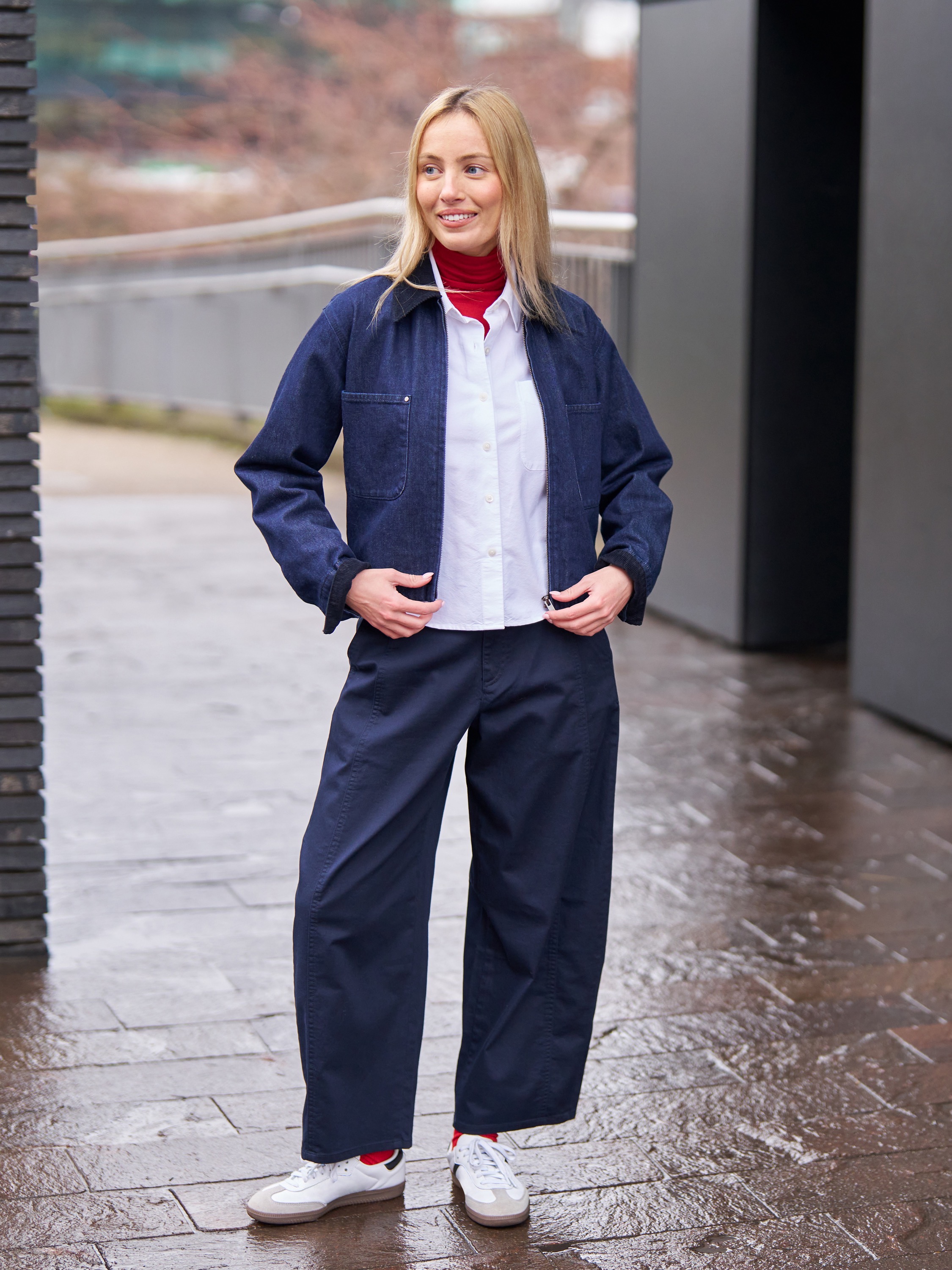Model draagt ​​een katoenen broek met curved pijpen, een rode coltrui, een witte blouse, een spijkerjasje, rode sokken en witte sneakers.